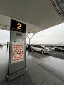 Info pillar at an airport terminal showing gate 2 and a No Smoking sign with cars parked nearby